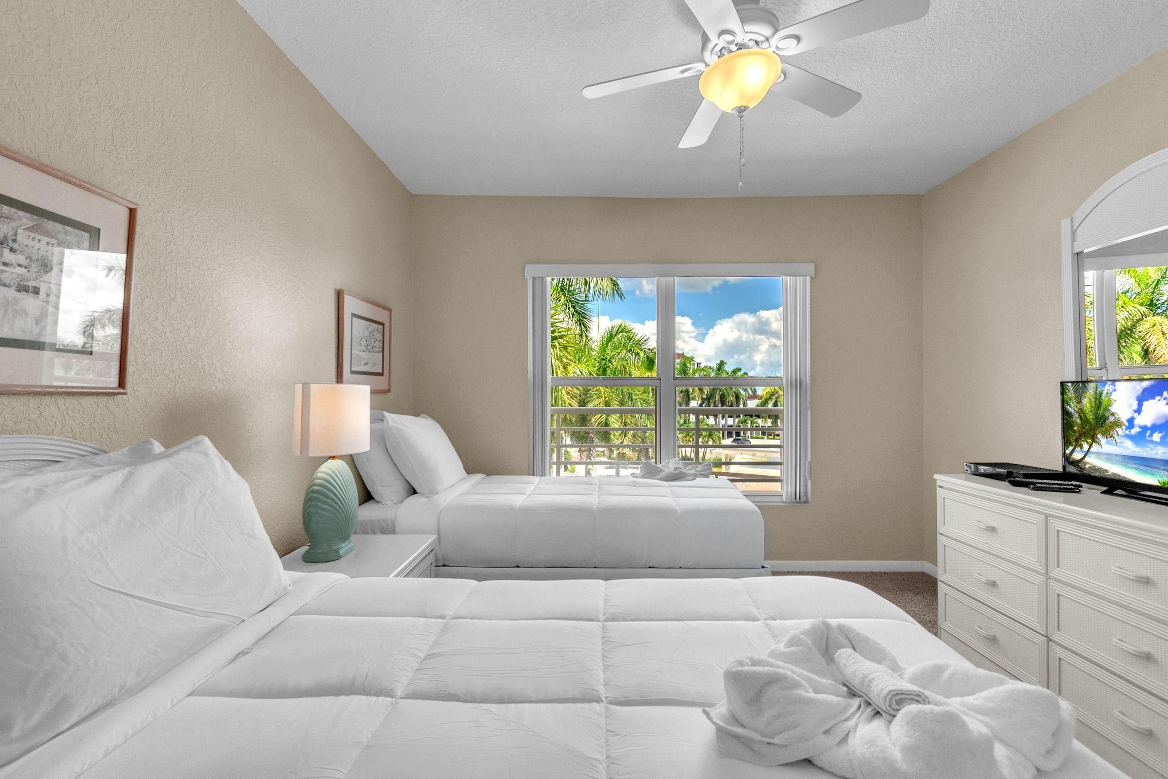 Habitación de hotel con dos camas, un ventilador de techo, una ventana con vistas a palmeras y un televisor sobre un tocador.