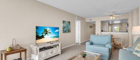 A living room with a television, a wicker coffee table, a blue sofa, and a kitchen area in the background.