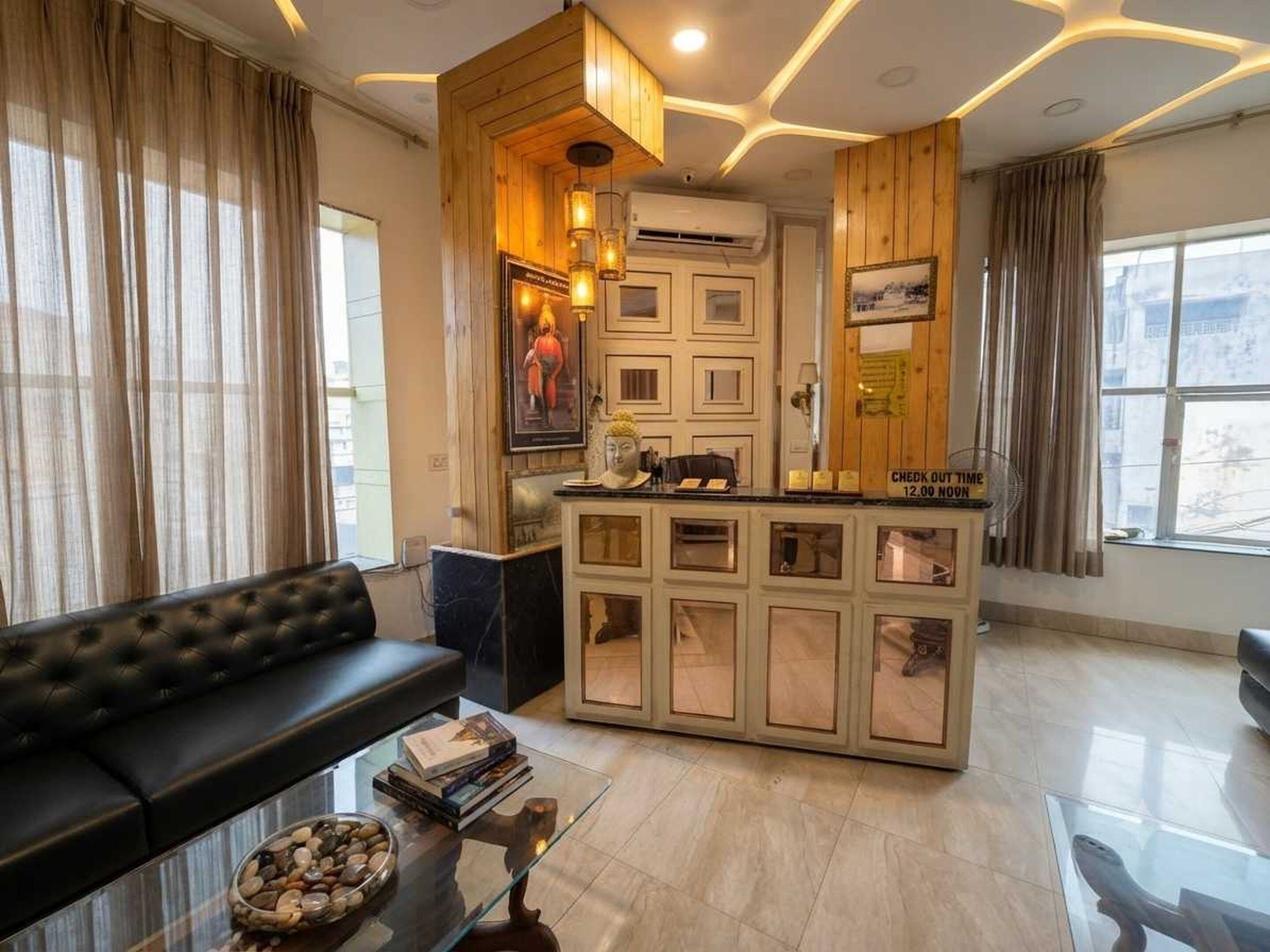 Een moderne hotellobby met een zwarte leren bank, een glazen salontafel met boeken en stenen, een receptiebalie met een uitcheck-bord en een grote Boeddha-beeld.
