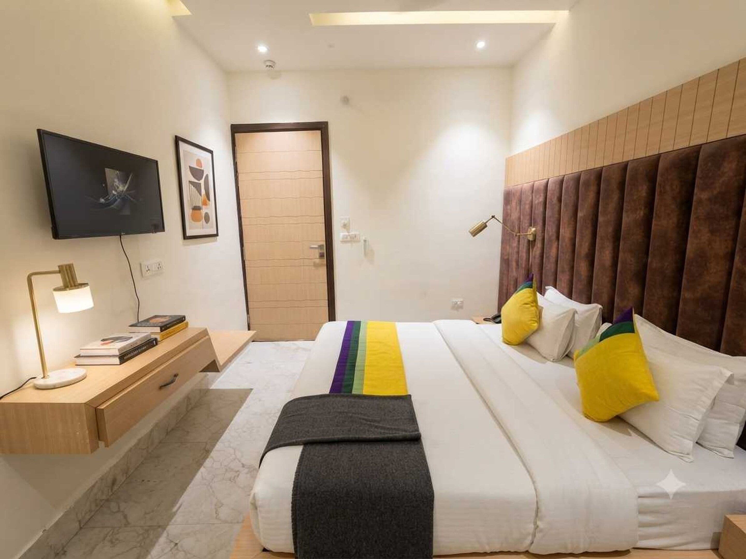 A modern hotel room with a bed, bedside table, lamp, and a TV on the wall.