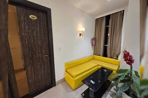 A room with a yellow sofa, a black coffee table, a wooden door with a number plate, a wall-mounted light, and a fire extinguisher. - Classic Sehdev Residency (Amritsar)