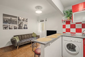 Un espace de vie compact comprenant un canapé, un lave-linge et un téléviseur fixé au mur.