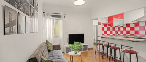 A compact living space with a sofa, a small table with a plant, a TV, and a kitchenette with red and white checkered backsplash.