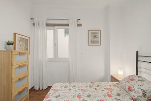 Une chambre à coucher avec un lit, une commode en bois, une fenêtre avec des rideaux, un tableau encadré au mur et une table de chevet avec une lampe.
