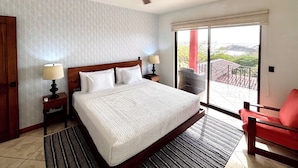 A hotel room with a large bed, two bedside tables with lamps, a ceiling fan, a red chair, and a view of the outdoors through a sliding glass door. - Villa Catalina 11 (Potrero)