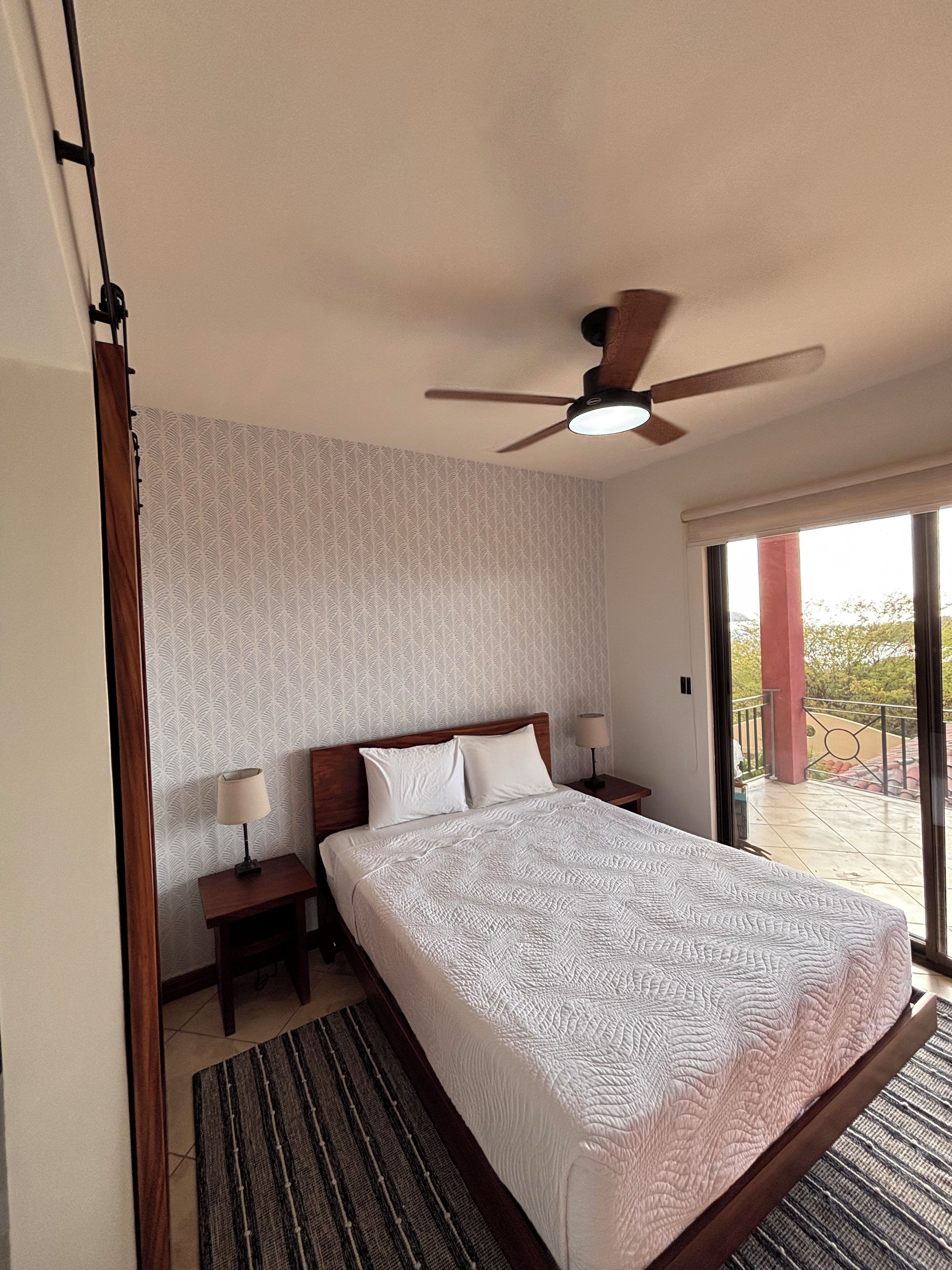 A bedroom with a bed, a ceiling fan, a nightstand with a lamp, a window with a view, and a patterned wall.
