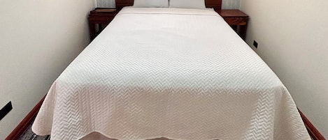 A neatly made bed in a room with a window, patterned carpet, and a nightstand with a lamp.