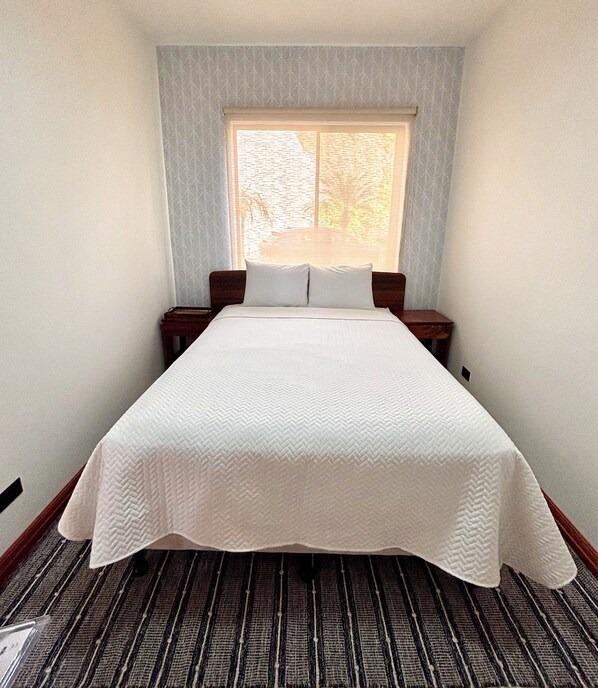 Una cama bien hecha en una habitación con ventana, alfombra estampada y mesita de noche con lámpara.