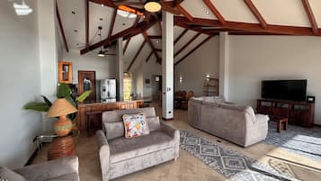 A spacious living area with a high ceiling, wooden beams, a sofa, a television, and a kitchen in the background.