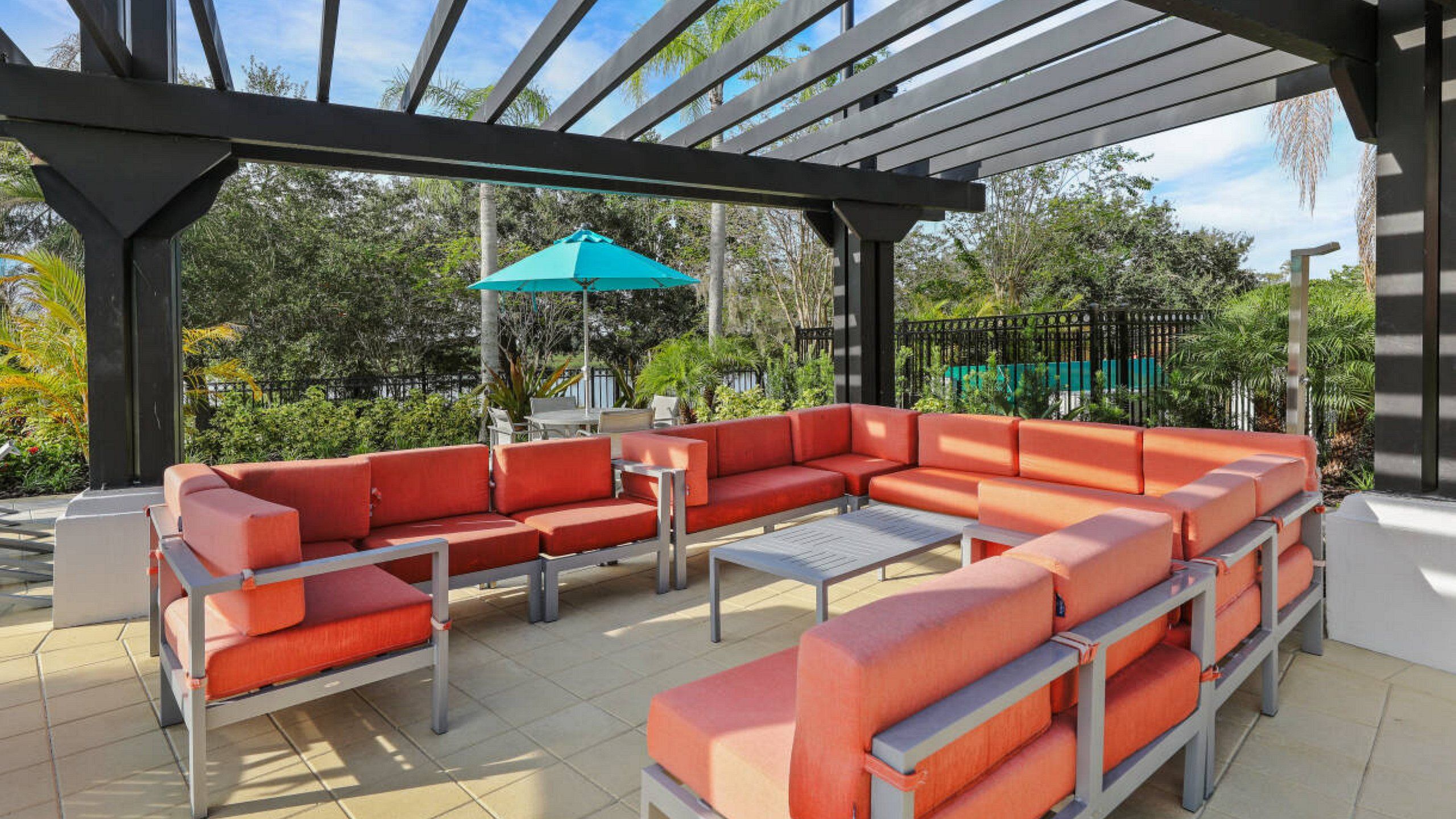 A covered outdoor seating area with a sectional sofa and a table, surrounded by greenery.