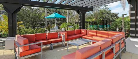 A covered outdoor seating area with a sectional sofa and a table, surrounded by greenery.