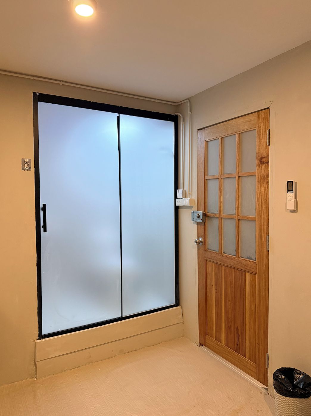 Una habitación con una puerta corredera, una puerta de madera con paneles de vidrio, un interruptor de luz y un cubo de basura.
