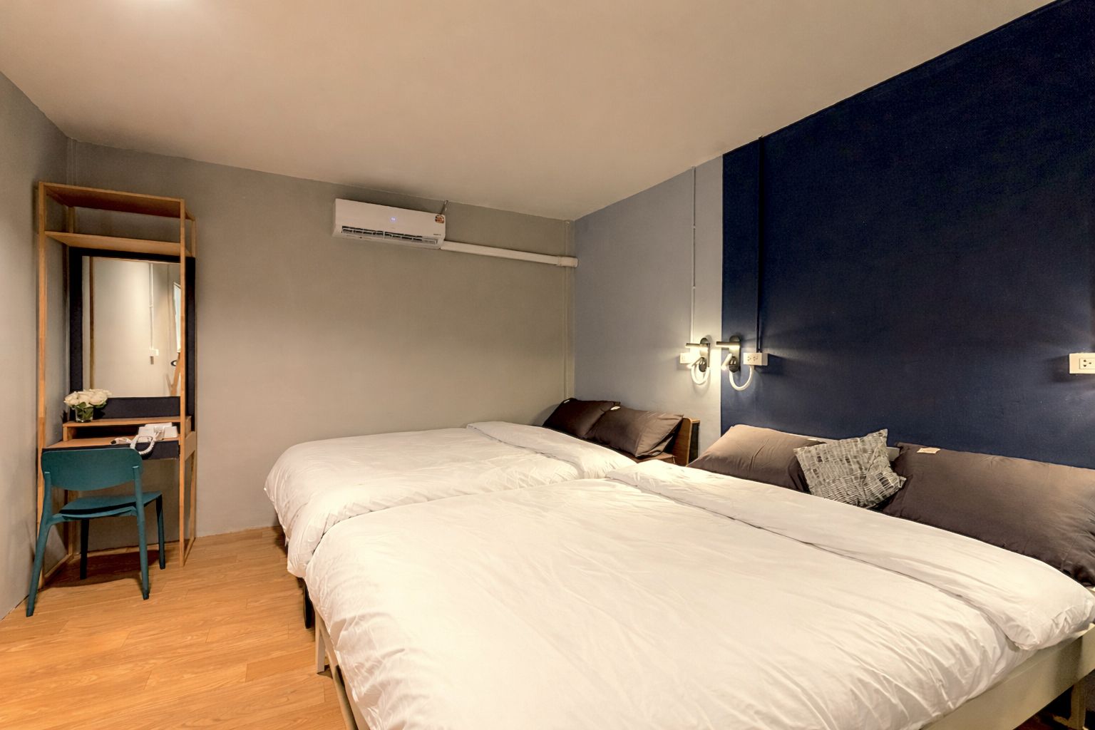 A bedroom with a large bed, a desk with a chair, a mirror, an air conditioning unit, and a wall-mounted light fixture.