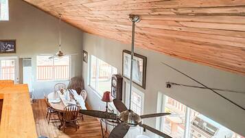 Um quarto amplo com teto alto, vigas de madeira, área de refeições com mesa e cadeiras, um sofá e um ventilador de teto.