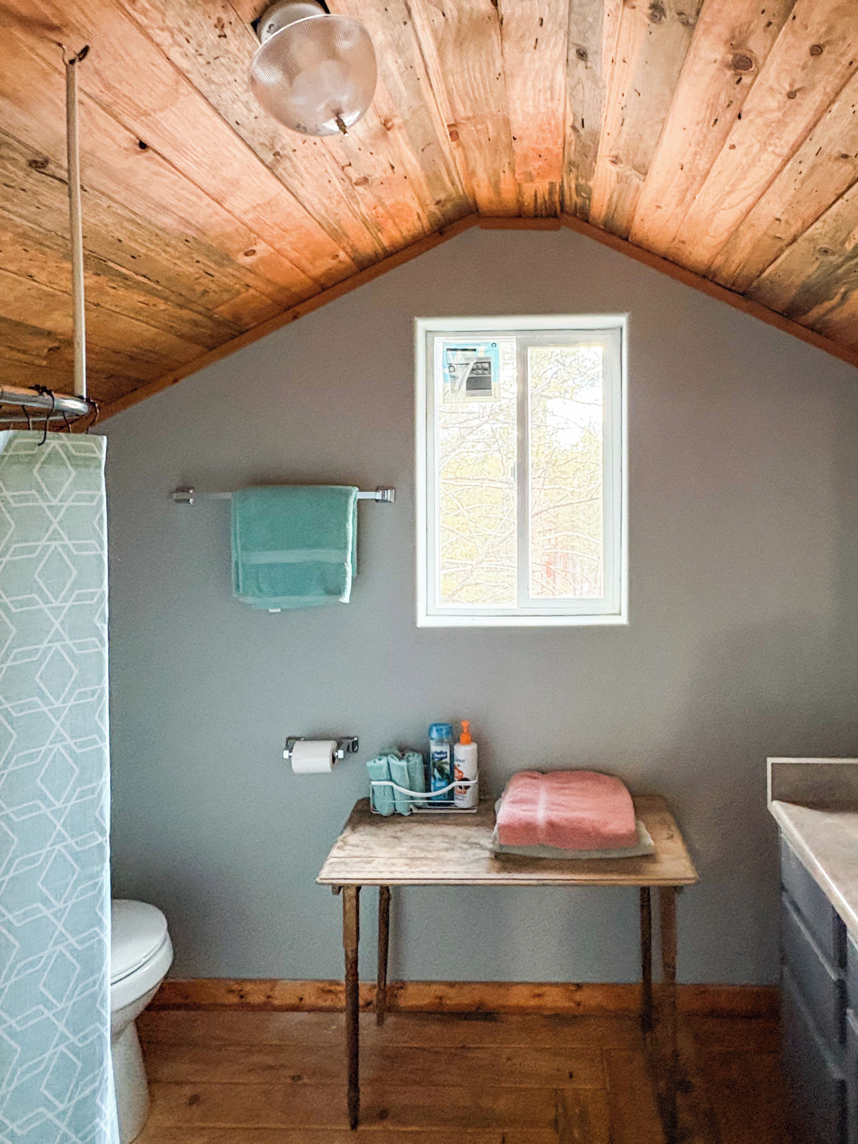A small bathroom with a wooden ceiling, a window, a towel rack, a toilet, and a table with toiletries.