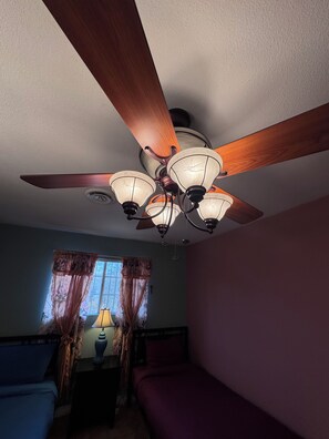 Une chambre avec un ventilateur de plafond, une fenêtre avec des rideaux, une lampe de chevet et un lit.