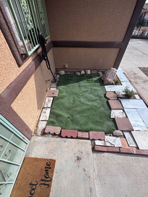 Un petit espace jardin aménagé avec du gazon synthétique, un paillasson portant l’inscription « Bienvenue chez vous » et différents types de pavés.
