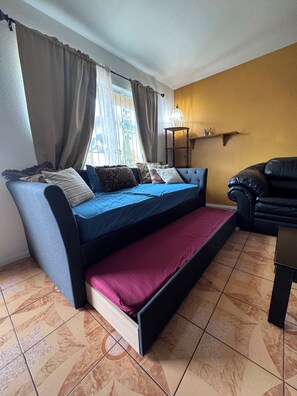 Quarto com sofá-cama, uma mesa pequena, luminária e janela com cortinas.