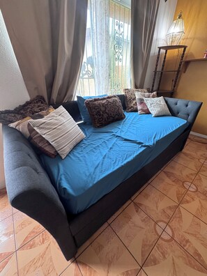 Un canapé-lit recouvert d’une housse bleue, agrémenté de plusieurs coussins, placé contre un mur près d’une fenêtre et de rideaux.