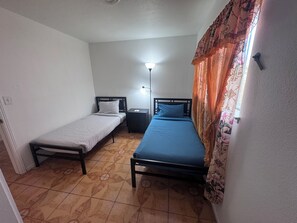 Acomodação pequena e individual com duas camas de solteiro, uma mesa de cabeceira, abajur e cortinas estampadas.