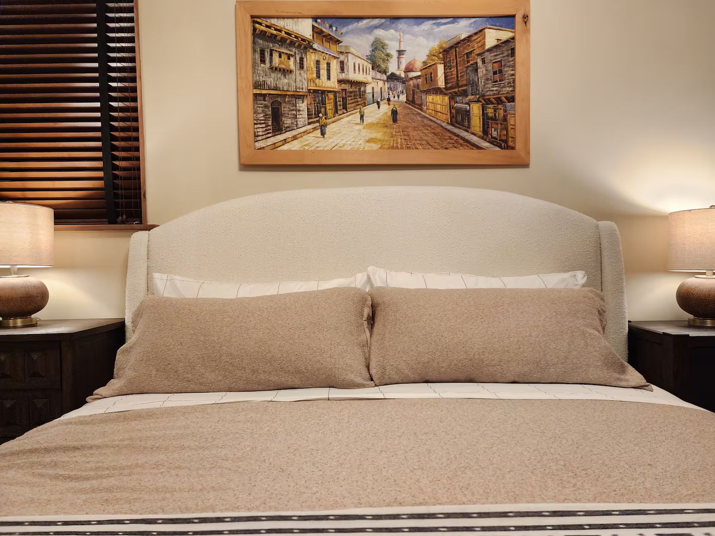 Uma cama com cabeceira, duas luminárias de apoio e um quadro na parede retratando uma cena de rua.