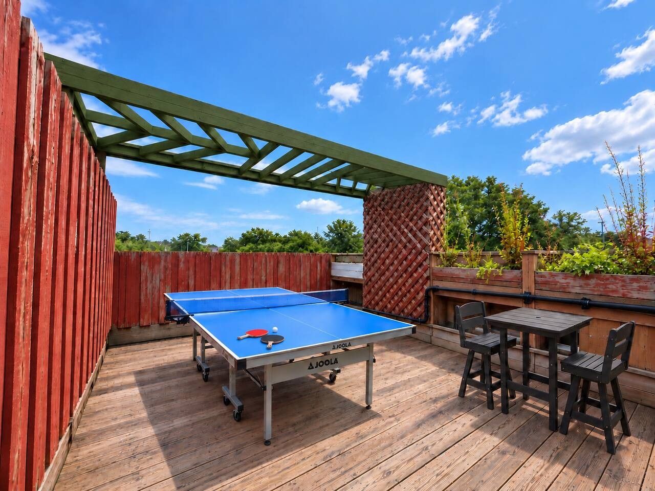 Une table de ping-pong installée à l’extérieur, avec deux chaises et une petite table, le tout posé sur une terrasse en bois, bordée d’une clôture rouge et surmontée d’une pergola verte.