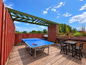 An outdoor ping pong table with two chairs and a table, set on a wooden deck with a red fence and a green pergola overhead.