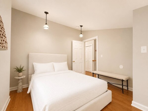 A bedroom with a large bed, bedside tables, a bench, and two pendant lights.