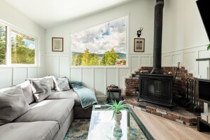 A living room with a sofa, a fireplace, a large window with a view of trees and hills, and a small table with a plant. - Park City Ski Getaway | Private Hot Tub | Fireplaces | Sleeps 10 (Park City)