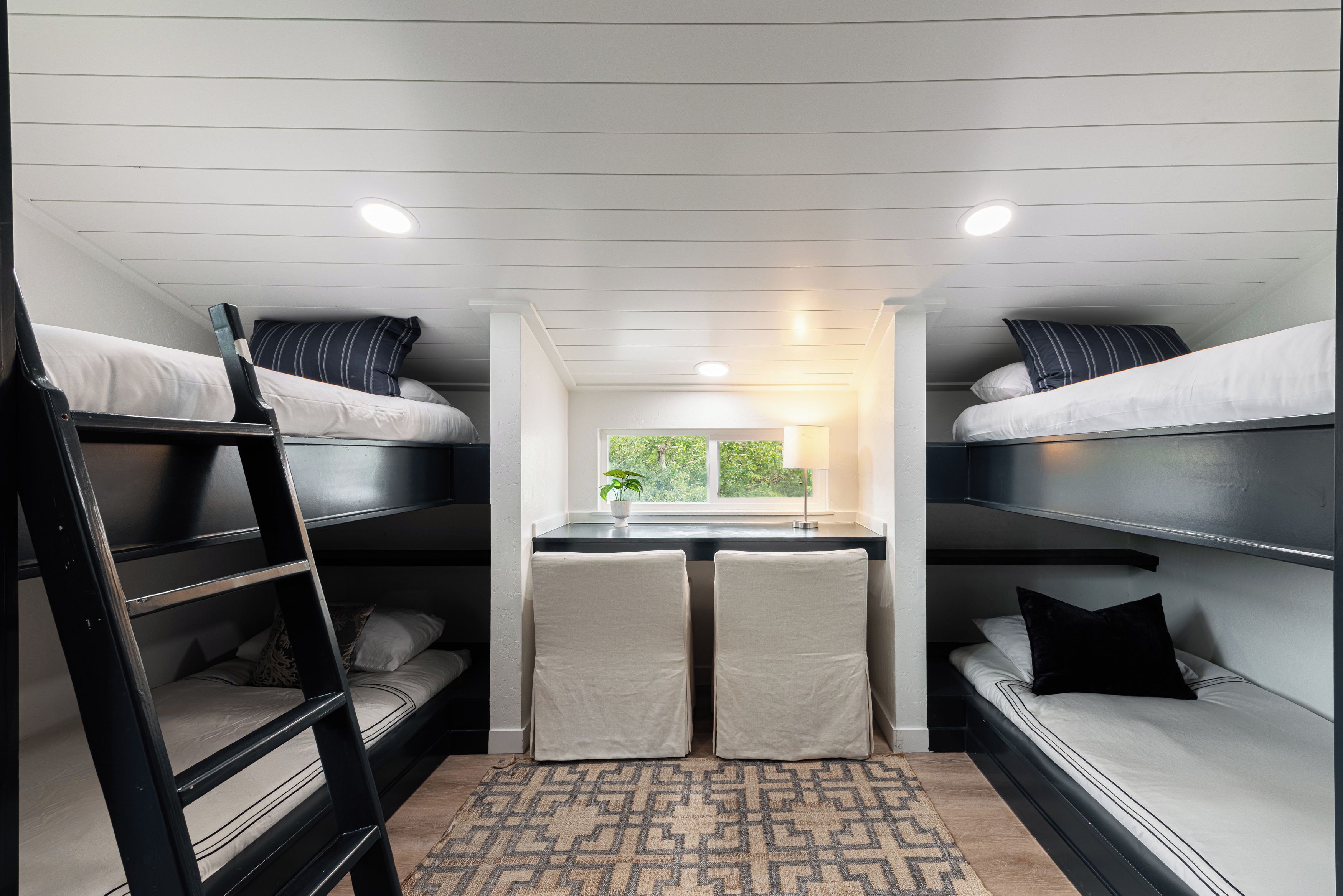 A compact, well-lit dormitory room with bunk beds, a small desk, and a window providing a view of greenery outside.