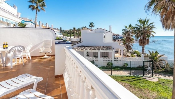 Un balcón con vista al mar, mobiliario exterior blanco y palmeras.