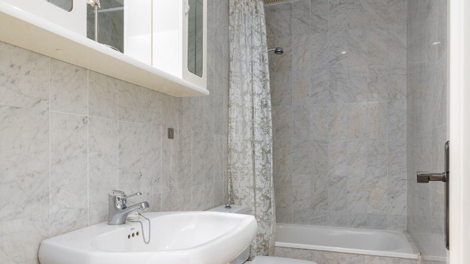 A bathroom with a white sink, a shower, and a marble-tiled wall.