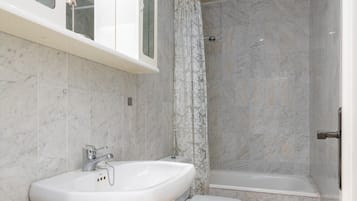 A bathroom with a white sink, a shower, and a marble-tiled wall.