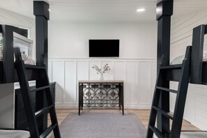Ein Zimmer mit weißer Wand, einem an der Wand montierten Flachbildfernseher, einem dekorativen Metall-Konsolentisch und Möbeln mit schwarzem Rahmen.