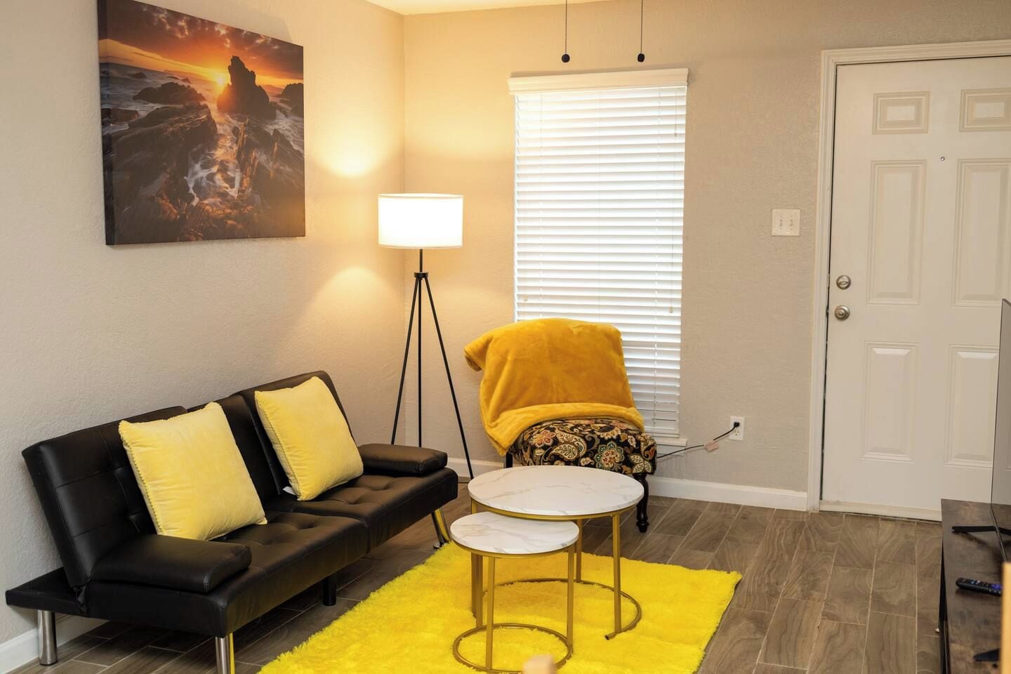 A living room with a black sofa, yellow cushions, a small round table, a floor lamp, and a window with blinds.