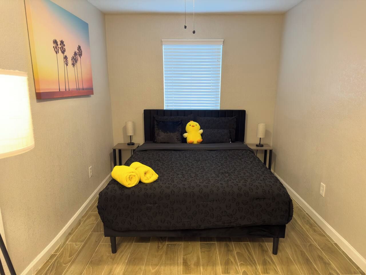 Une chambre à coucher comprenant un lit, deux tables de chevet, une fenêtre avec des stores et un tableau accroché au mur.