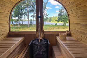 A sauna with a view of a lake and trees, wooden benches, and a metal stove. - Schweden-Auszeit: Lake Lodge mit Wintergarten, Sauna, Kamin & Boot direkt am See (Alsterbro ( Schweden))