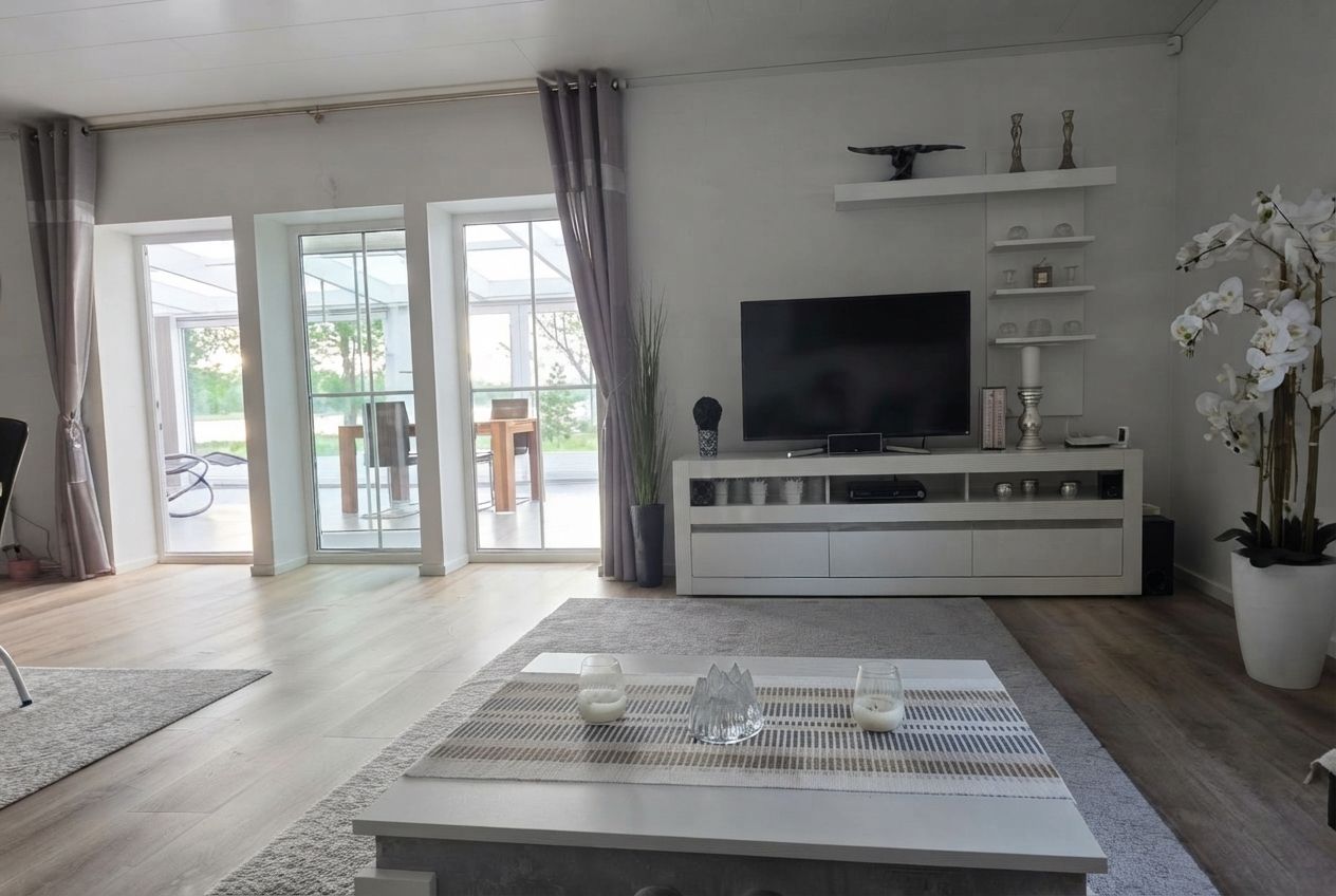 Una sala moderna con un televisor de pantalla plana, un mueble blanco para entretenimiento y un gran ventanal con cortinas.