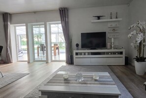 A modern living room with a flat-screen TV, a white entertainment unit, and a large window with curtains. - Schweden-Auszeit: Lake Lodge mit Wintergarten, Sauna, Kamin & Boot direkt am See (Alsterbro ( Schweden))
