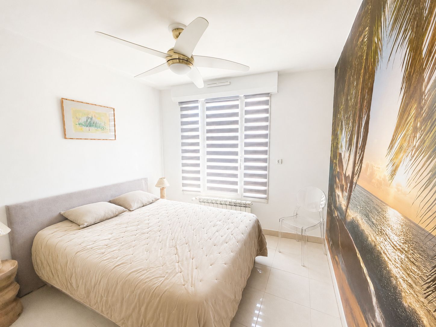 A bedroom with a bed, a ceiling fan, a window with blinds, a radiator, and a framed picture on the wall.