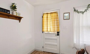 Une pièce avec une porte blanche, un rideau à carreaux jaunes, une étagère en bois avec une plante et un tableau encadré au mur.