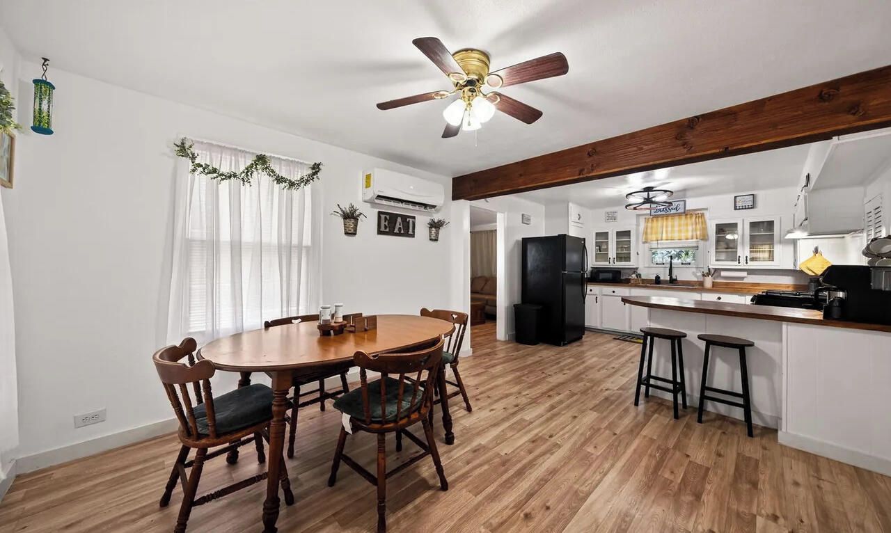 A dining area with a wooden table and chairs, a kitchen with a refrigerator and bar stools, and a ceiling fan.