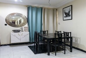 A dining area with a marble floor, a white cabinet with gold handles, a black dining table with chairs, a blue curtain, and a framed picture on the wall. - A Downtown Gbetsile Studio Balcony + King Bed (Tema)