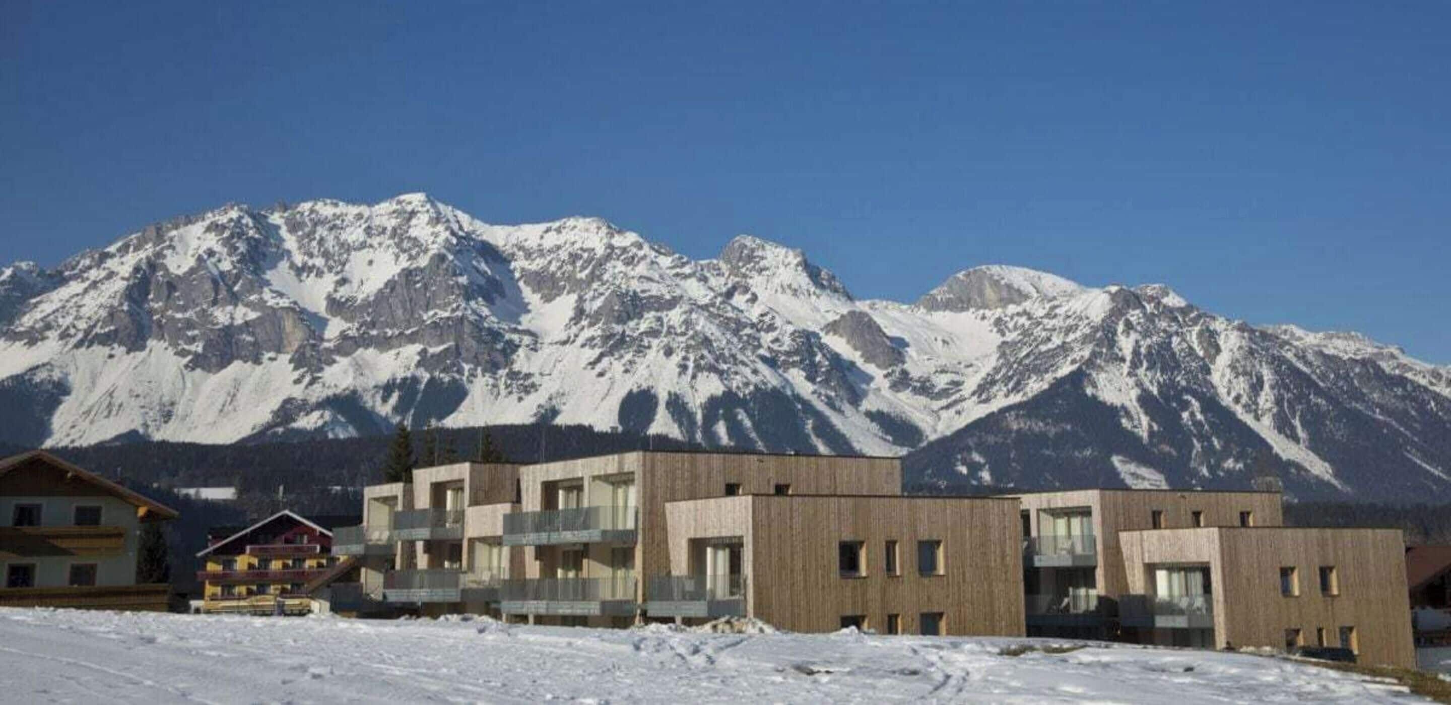 A modern building with a flat roof and a balcony, situated in a snowy landscape with mountains in the background.