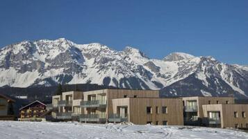 A modern building with a flat roof and a balcony, situated in a snowy landscape with mountains in the background.