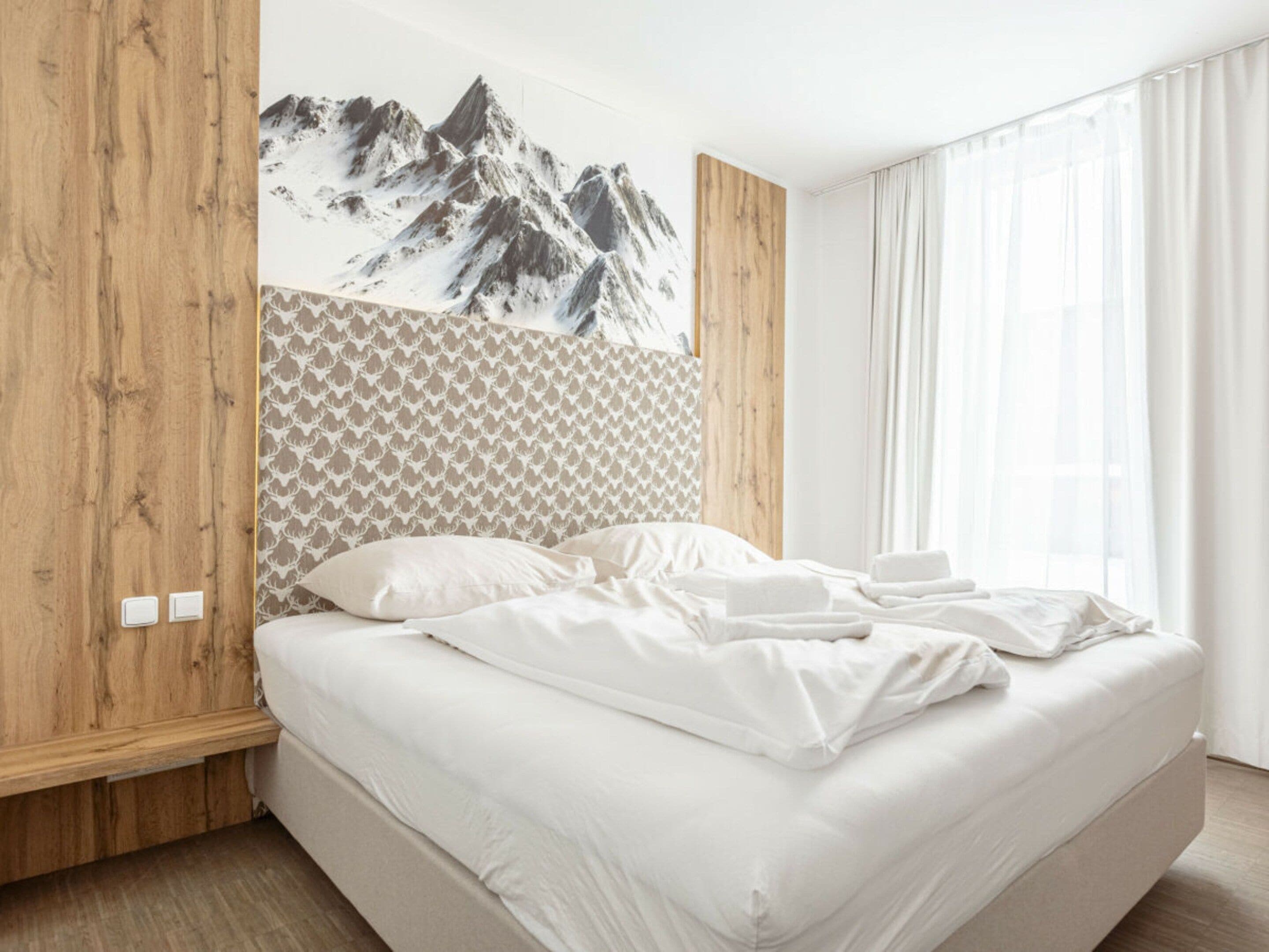 Un dormitorio moderno con cama, paneles de madera, una obra de arte con temática de montaña y cortinas traslúcidas.