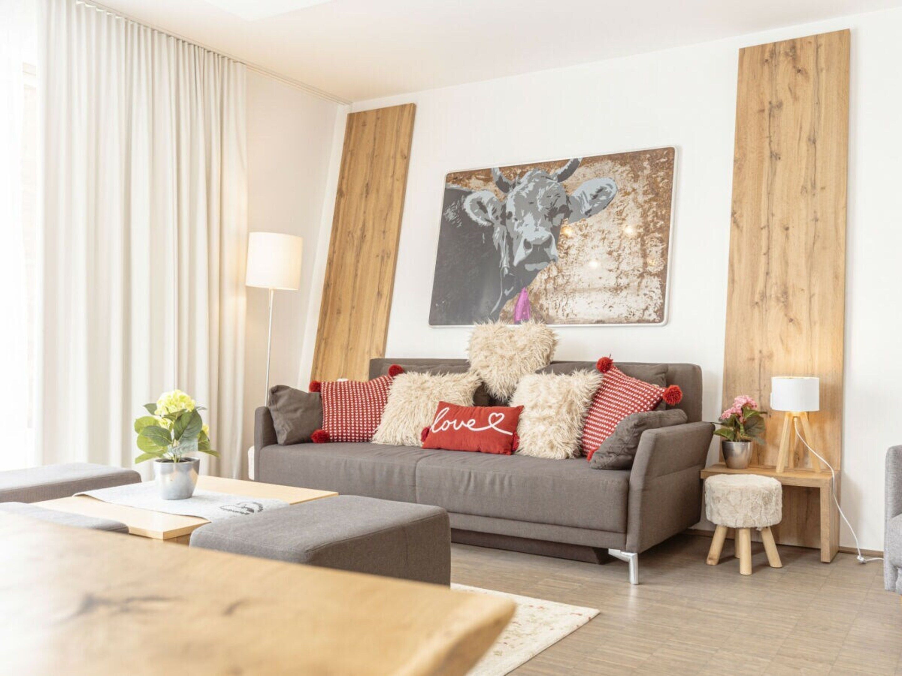Ein modernes Wohnzimmer mit einem grauen Sofa, roten und weißen Kissen, einem Holztisch und einem Bild einer Kuh an der Wand.