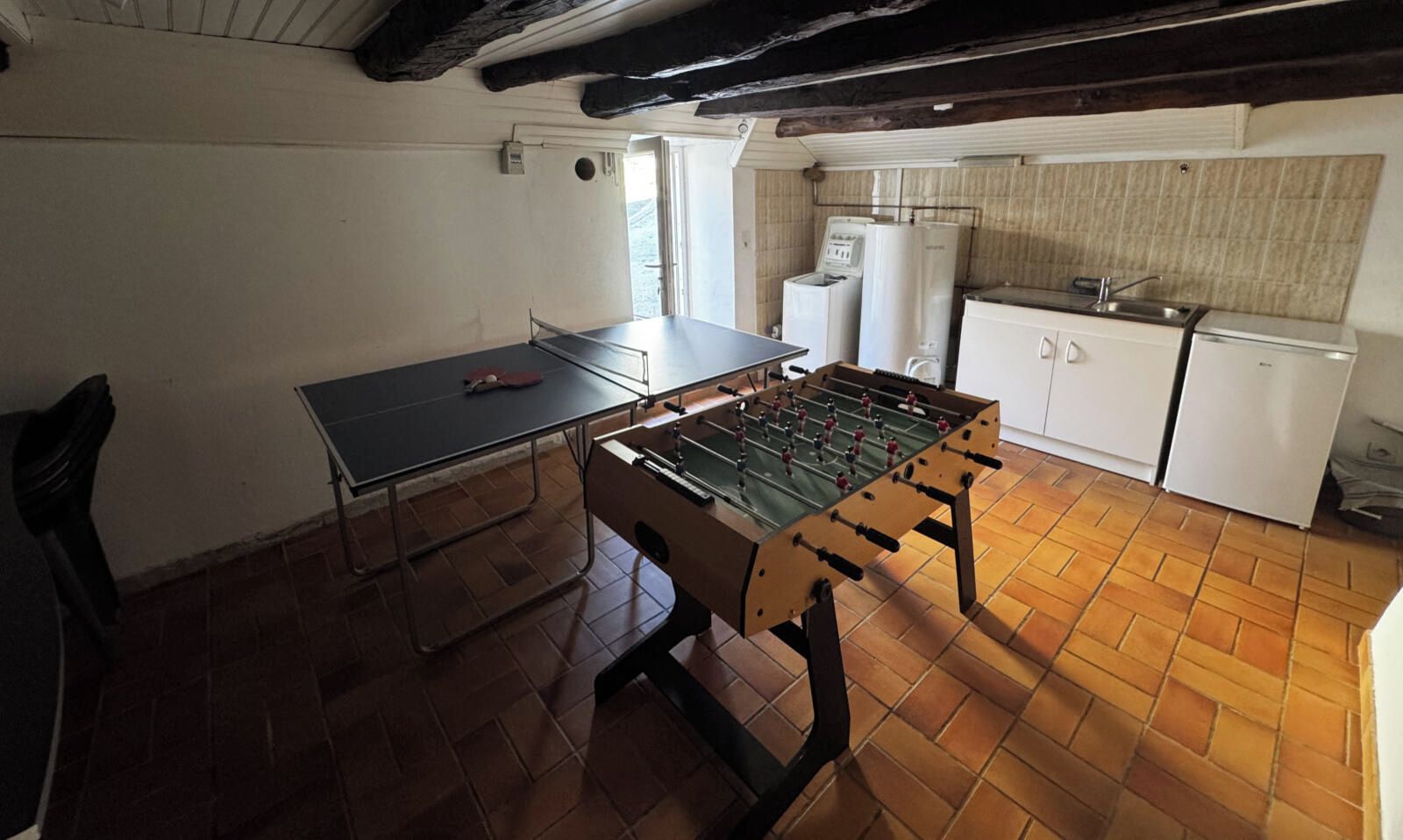 A room with a foosball table, a ping pong table, and a kitchenette area.