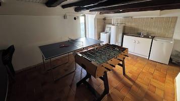 A room with a foosball table, a ping pong table, and a kitchenette area.