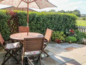 An outdoor patio with a round wooden table and chairs, a floral umbrella, and a hedge in the background. - The Croft (Ashbourne)